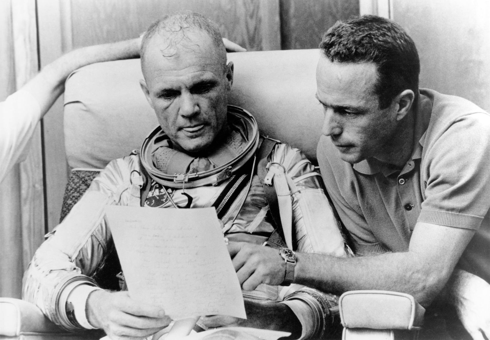 Astronauts John Glenn and Scott Carpenter review notes after Glenn's mission simulation run
