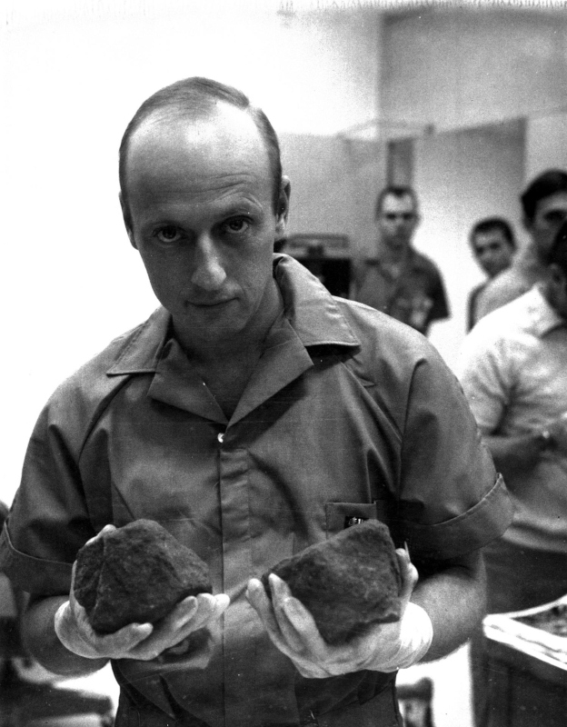 In white-gloved hands, Conrad shows two large moon rocks to the camera, as Lunar Receiving Laboratory technicians look on in the background