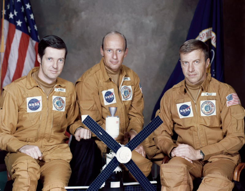 Official portrait of the Skylab 2 crew of Joe Kerwin, Conrad and Paul Weitz seated around a model of the space station. The real thing would not look so pristine when they reached it on orbit