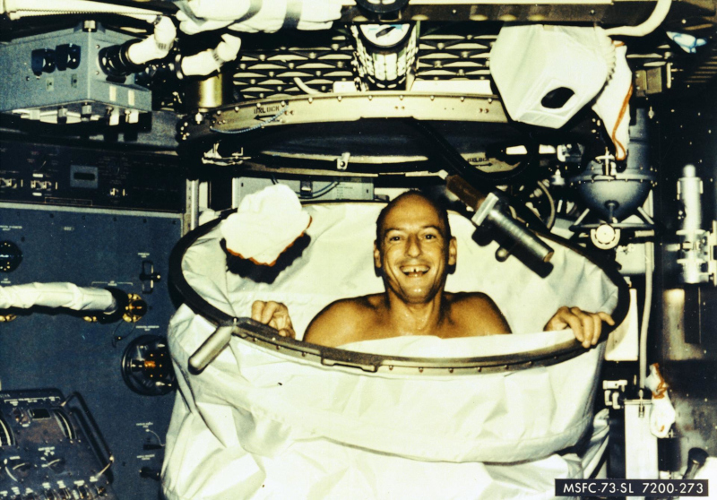 A laughing Pete Conrad looks at the camera from inside the Skylab shower, a white tubular structure that was attached to the ceiling of the space station to contain the water