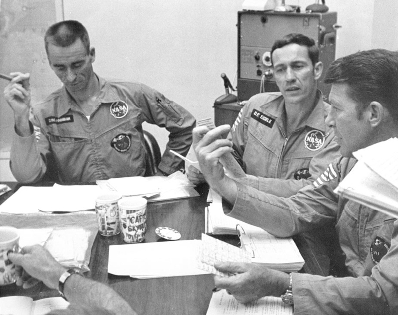 a black and white image of the Apollo 7 crew seated at a desk during a debriefing after the mission