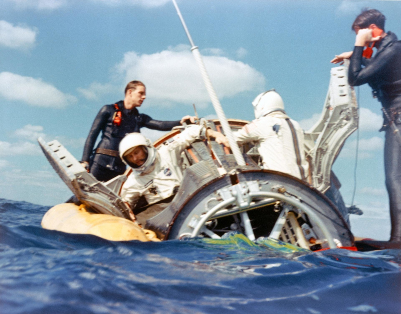 Navy divers stand on the floatation collar to assist the Gemini 6 crew in opeining the spacecraft hatches after splashdown.