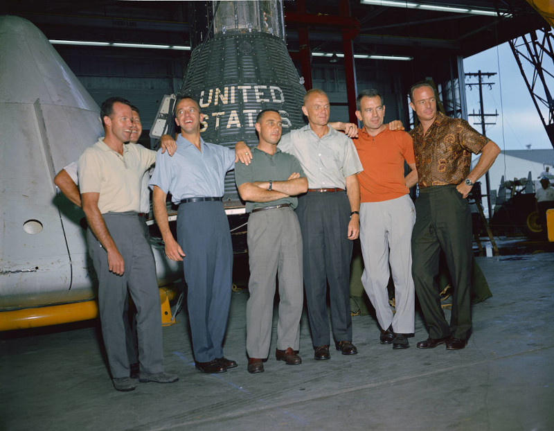 group photo of the original seven NASA astronauts informally dressed standing in a hangar in front of a spacecraft