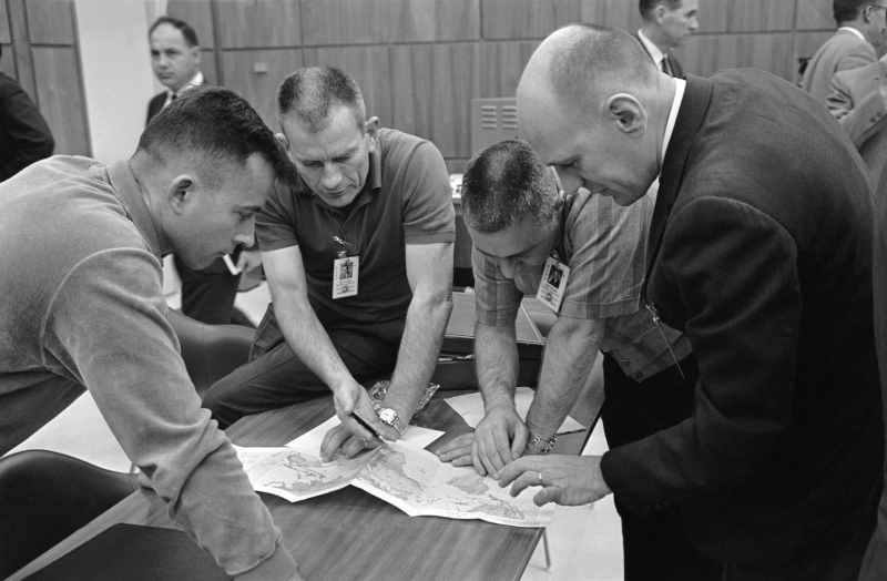 The crew of Gemini 3 gather around a table to examine a map of their planned orbital track with Chief Astronaut Slayton