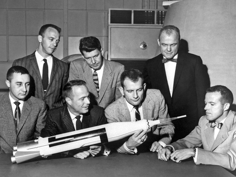 the original seven NASA astronauts are seen examining a model of the Atlas launch vehicle, with Slayton holding the model up for inspection
